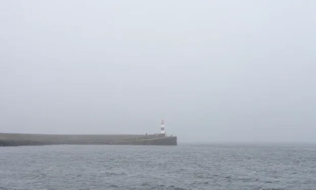 Thick coastal fog swallowing a harbour, a beacon reduced to a pale smear above flat grey water.