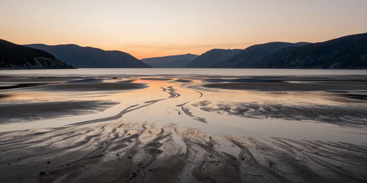 Tidal sand flats at first light, mirroring a pale orange sky across a fjord