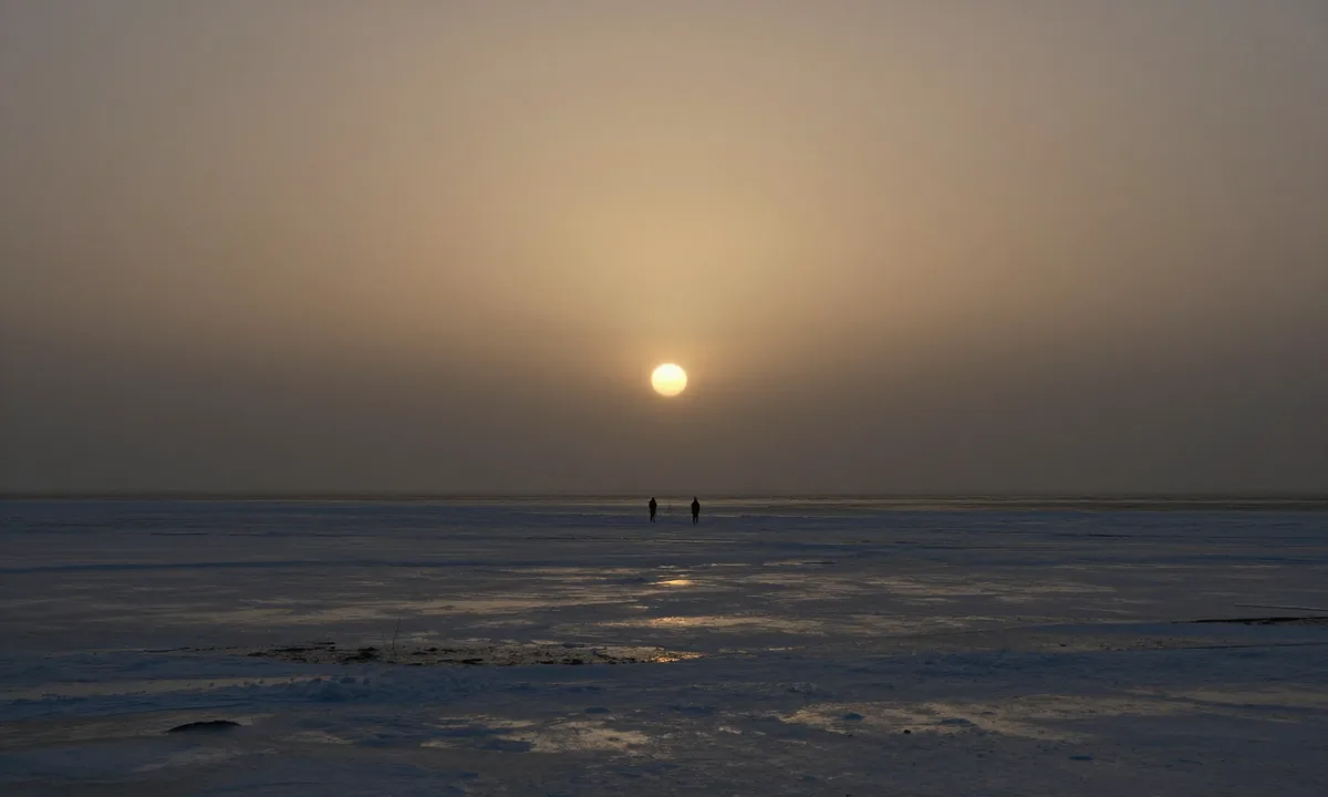 Two distant figures on a frozen northern shore beneath a low sun burning through sea haze