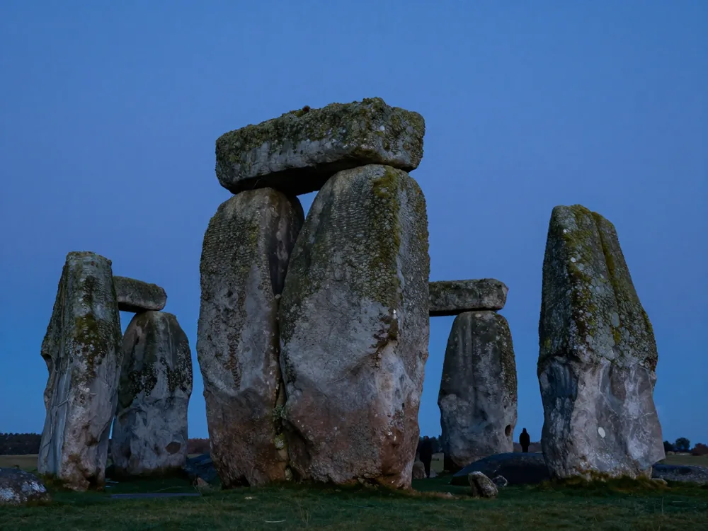 A weathered sarsen trilithon against a deep twilight sky, one distant figure at its base for scale