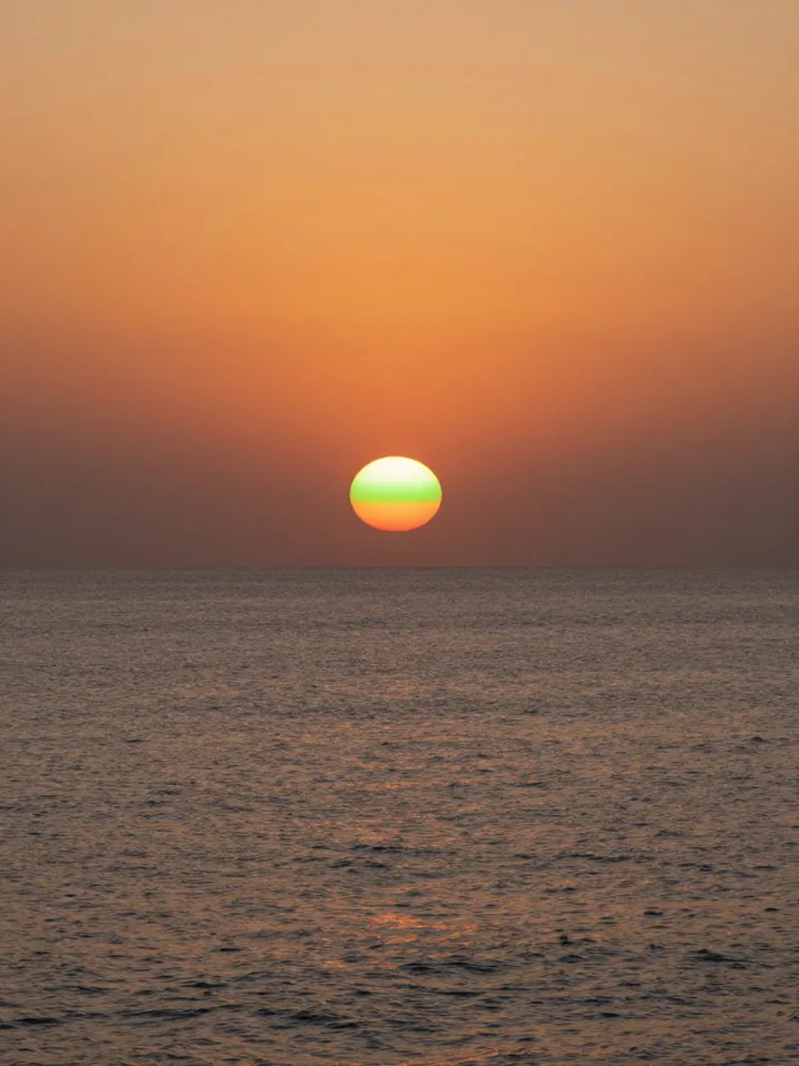 A setting sun crowned by a thin green rim over a calm sea horizon