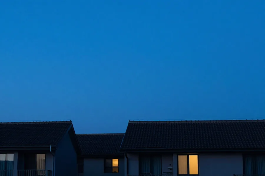 An even cobalt sky over dark rooftops, with two warm windows lit against it.