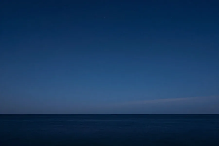 A faint pale horizon line under a deep indigo sky, the first stars showing over calm water.
